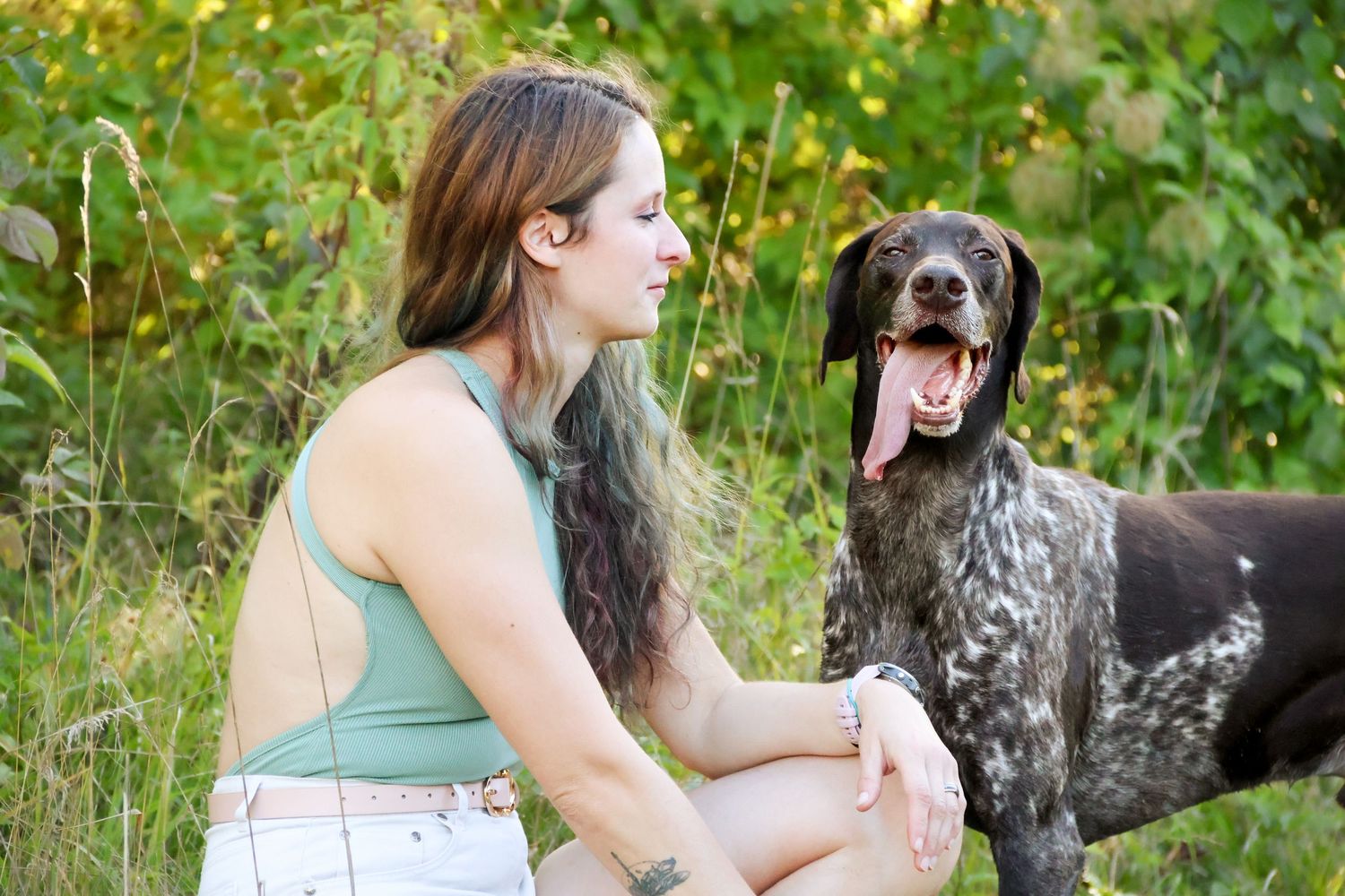 A young woman with long brown hair sits beside her spotted dog in a lush green outdoor setting.