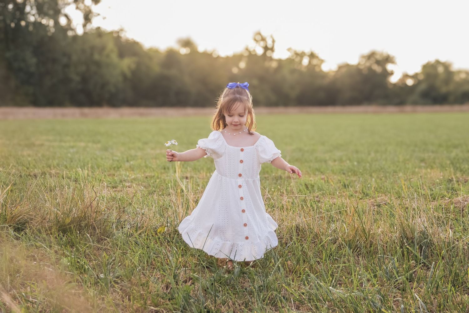 Little girl in white dress twirling in a green meadow holding a flower at sunset.