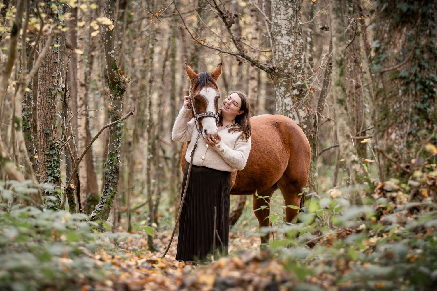 A woman in a cream sweater embraces a chestnut horse with a white blaze in an autumn forest.