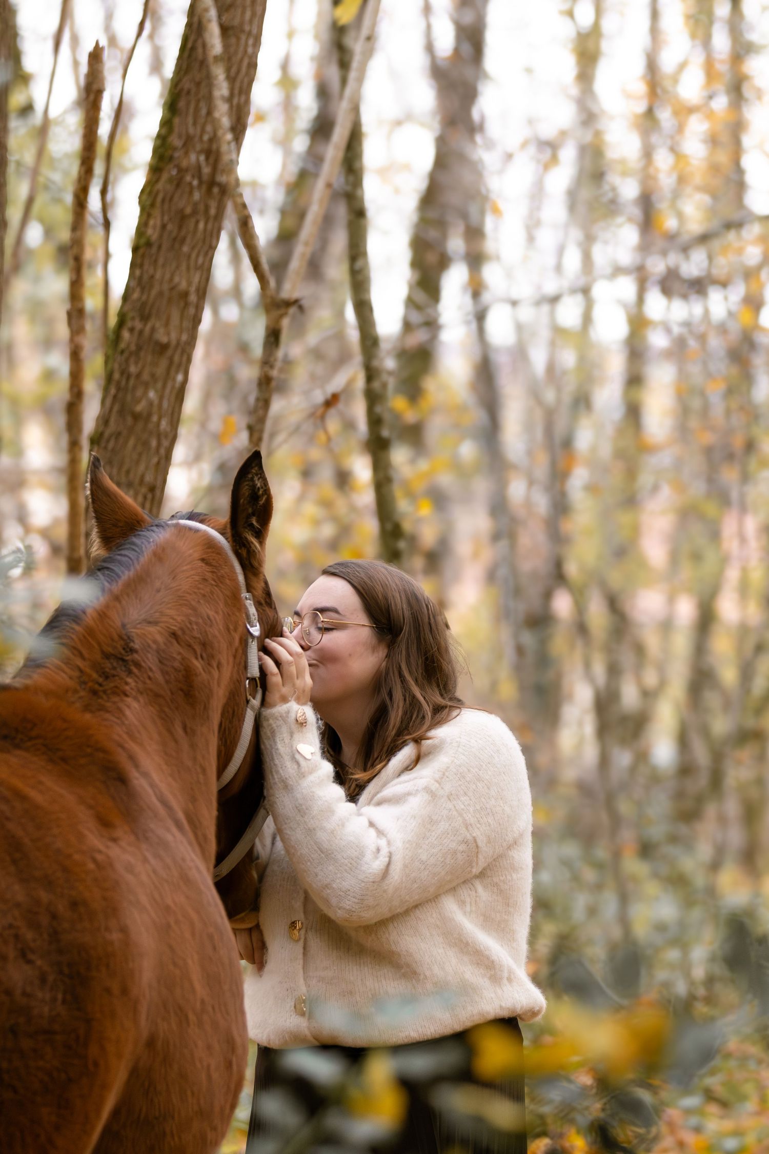 Woman in cream sweater kissing a chestnut horse's nose in an autumn forest setting.