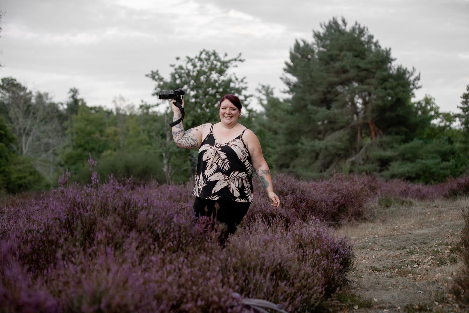 Femme souriante tenant un appareil photo en se promenant dans des champs de bruyère violette en extérieur.