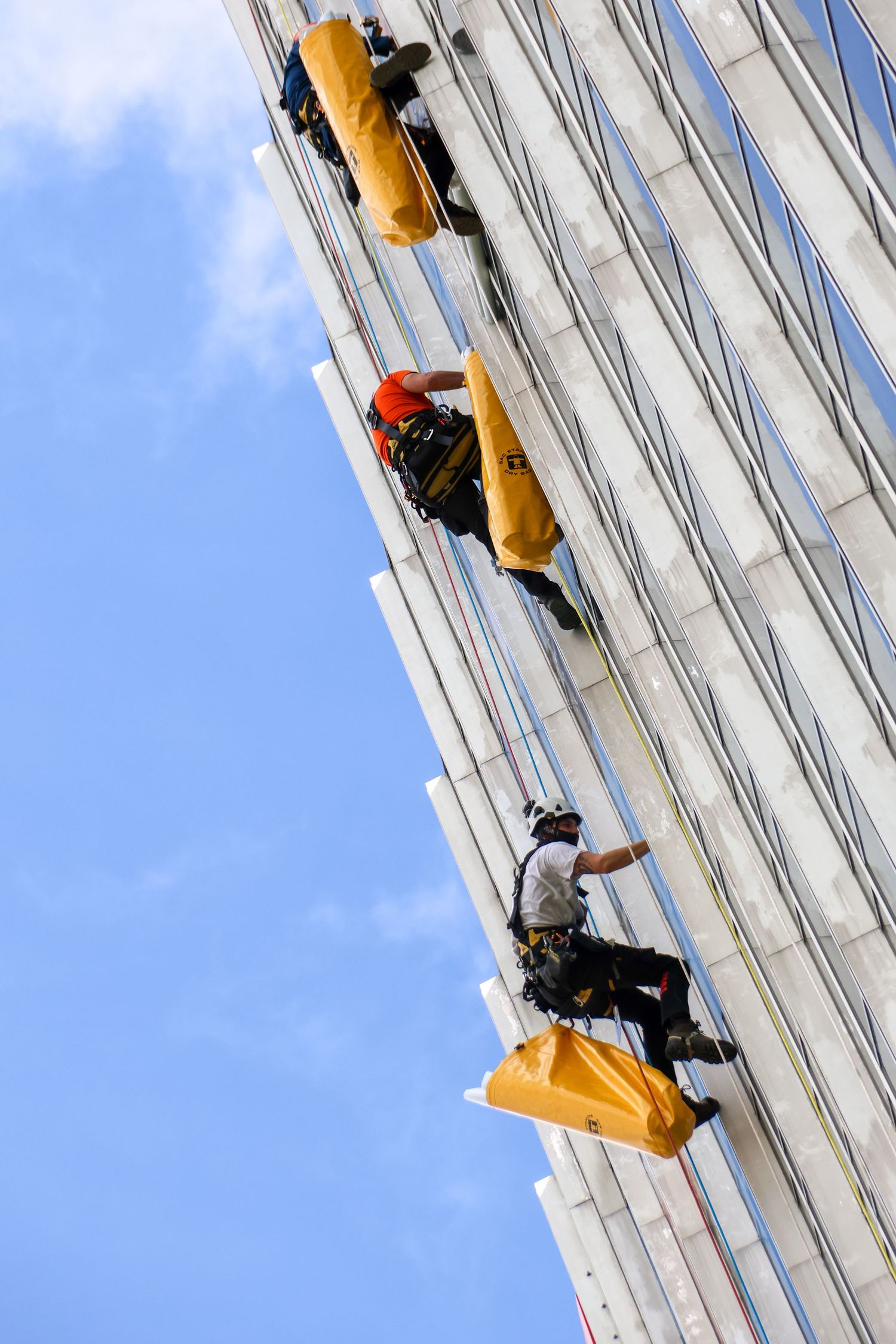 Three window washers rappel down a tall glass skyscraper facade using ropes and yellow equipment under a blue sky.