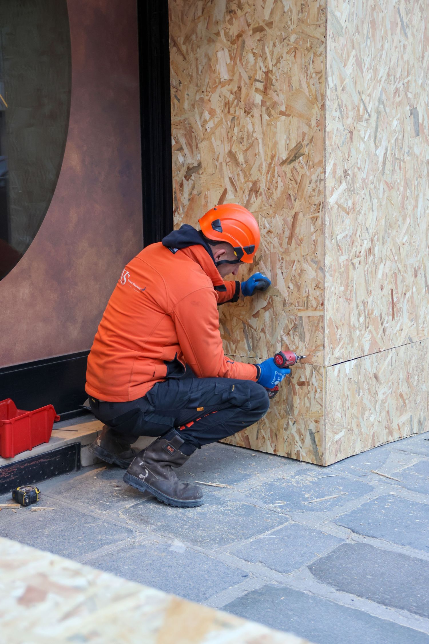 Construction worker in orange safety gear using a drill on a plywood-covered building exterior on cobblestone street.