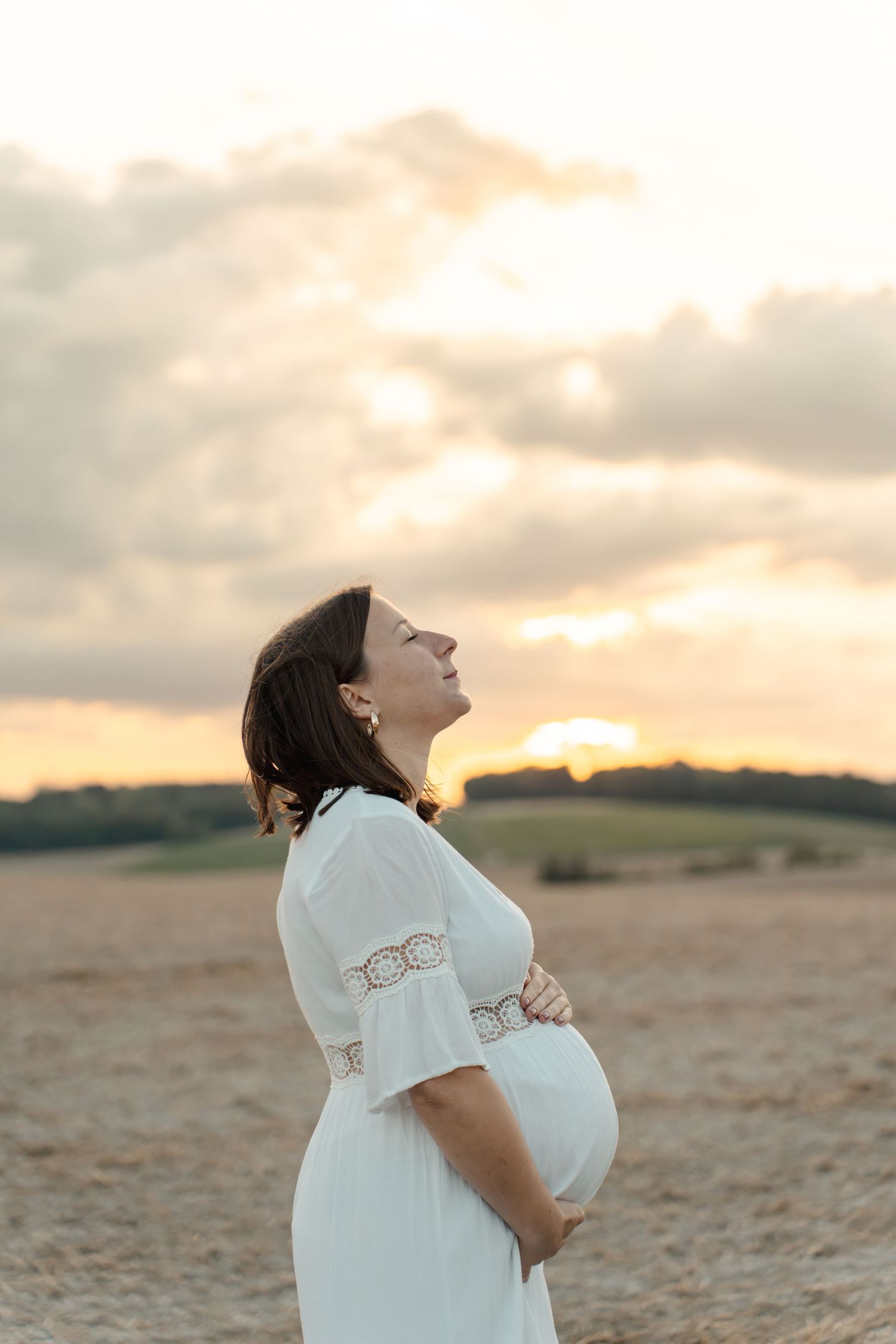 Pregnant woman in white dress holding baby bump at sunset in a golden field, eyes closed, smiling peacefully.