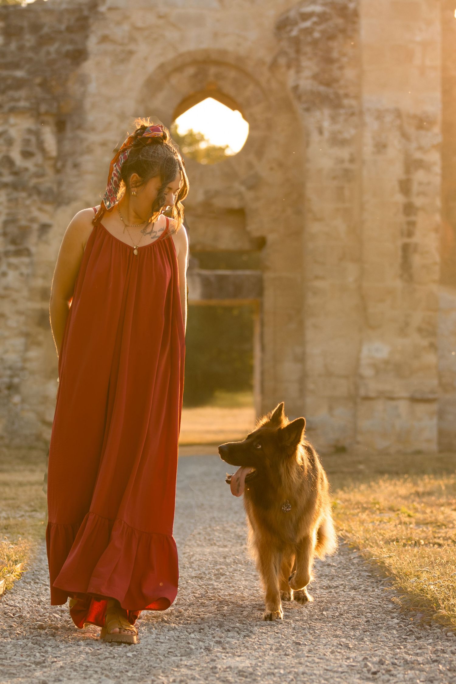 Woman in red dress walking with German Shepherd dog near ancient stone ruins at golden sunset.