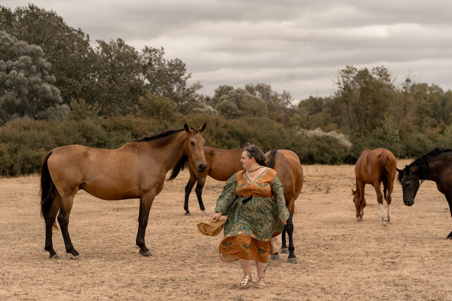 Woman in green dress feeding brown horses on a dry sandy field with trees in the background.