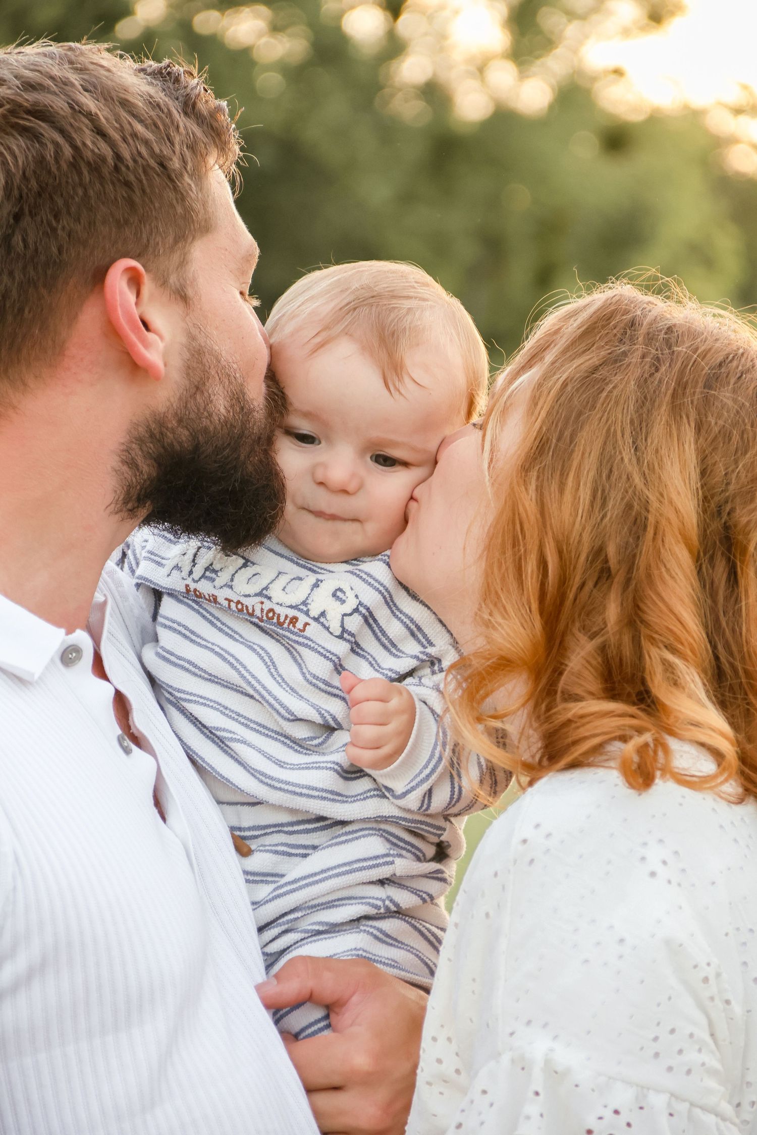 Loving parents kissing their baby on the cheeks simultaneously in a sunny outdoor setting.