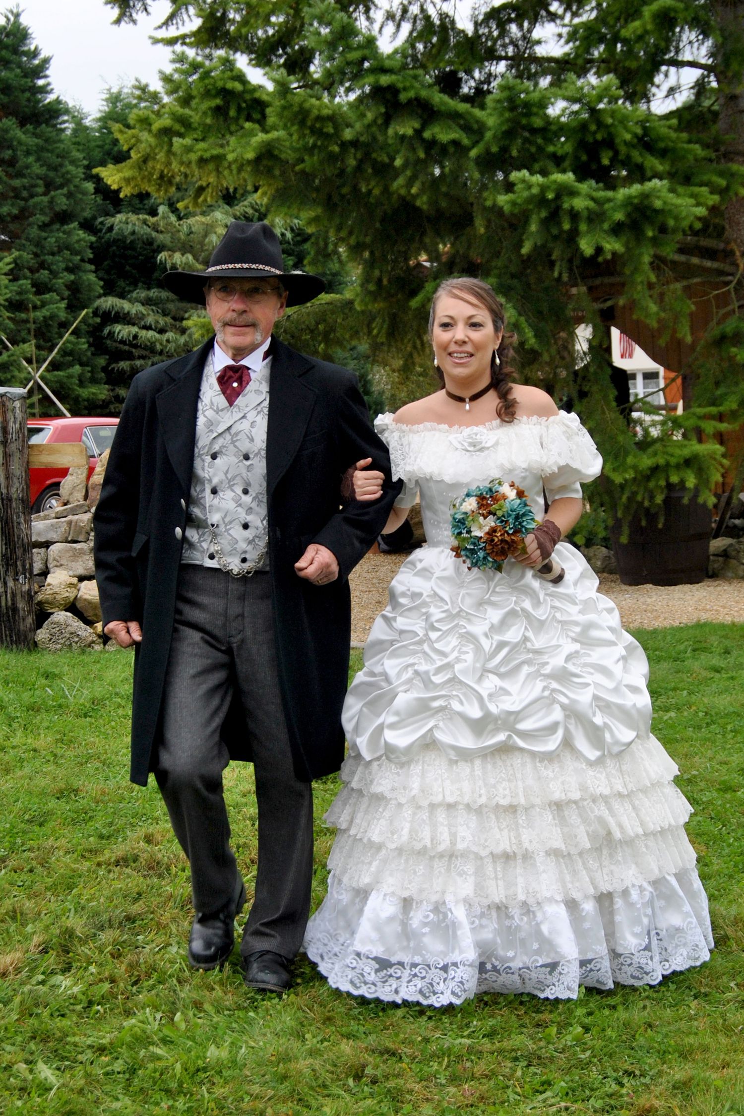 Bride in a ruffled white gown walks arm-in-arm with a man in a black cowboy hat and long coat at a western wedding.