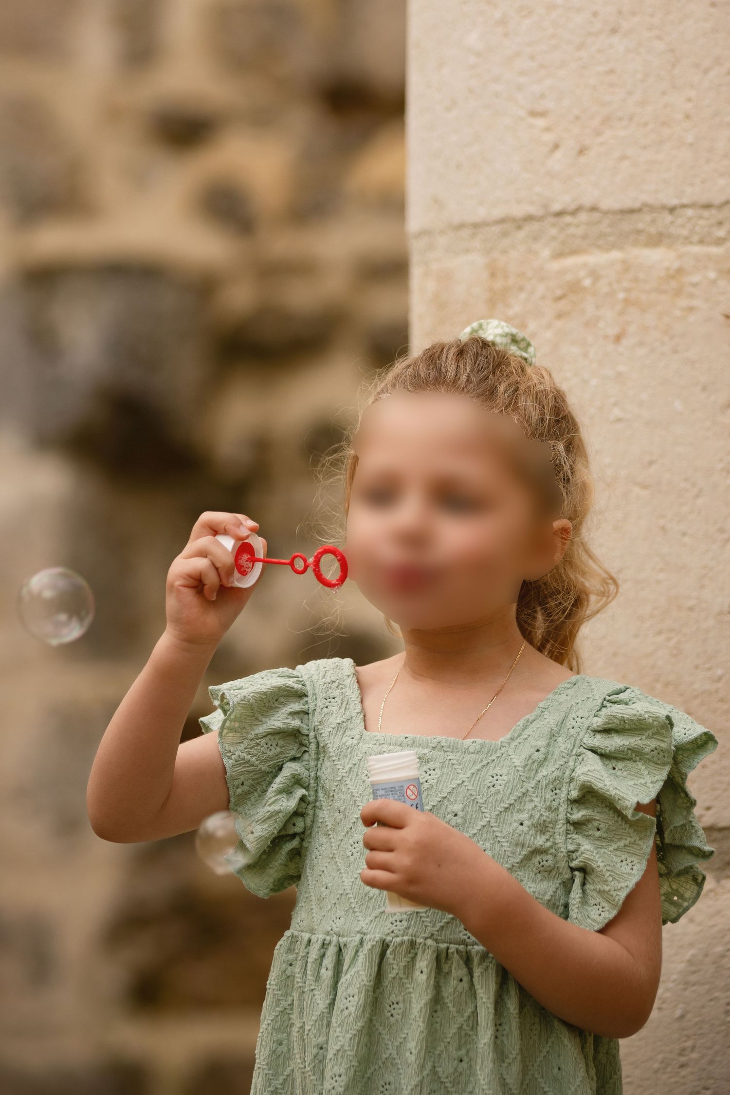 Young girl in green dress blowing soap bubbles outdoors near stone wall.