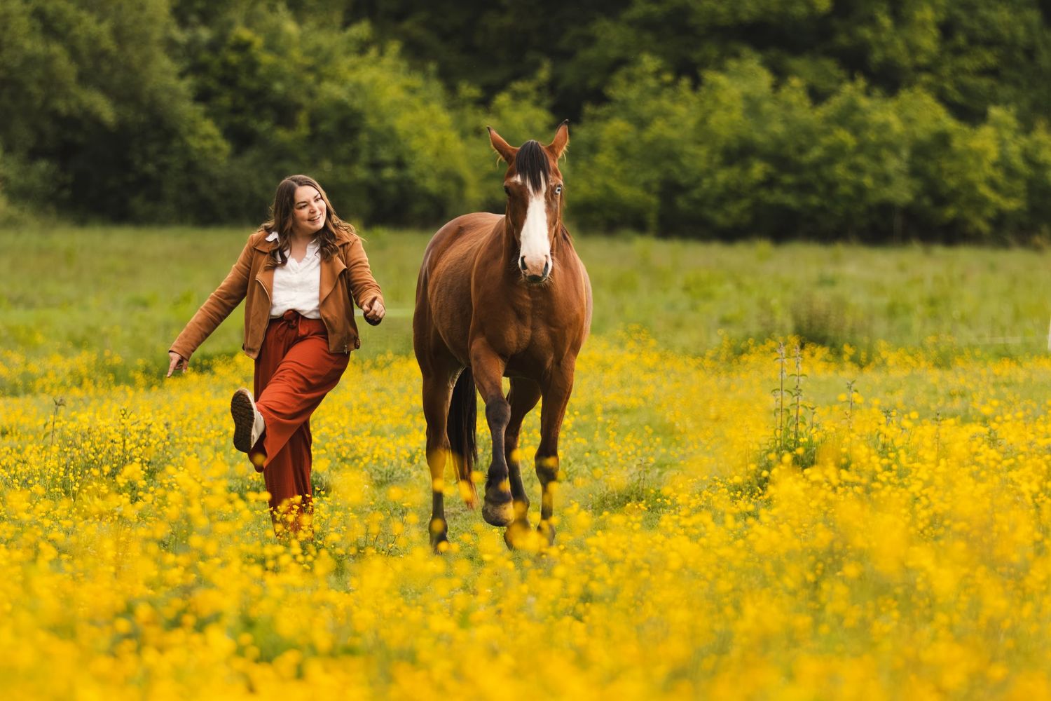 Woman in brown outfit running with a chestnut horse through a vibrant yellow wildflower meadow.