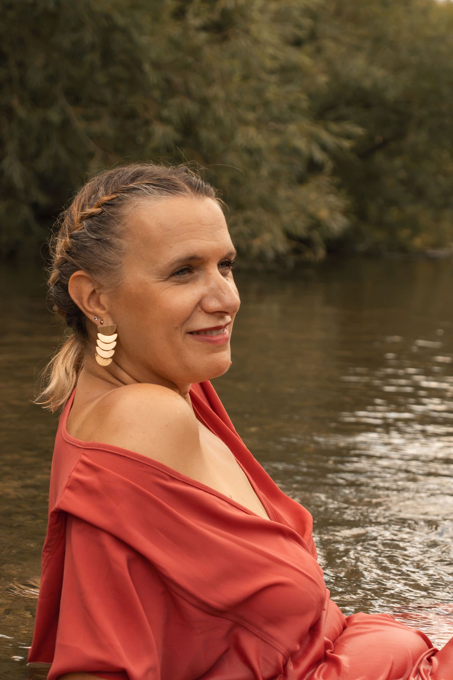 A smiling middle-aged woman in a red off-shoulder dress with gold earrings poses near a river with green trees.