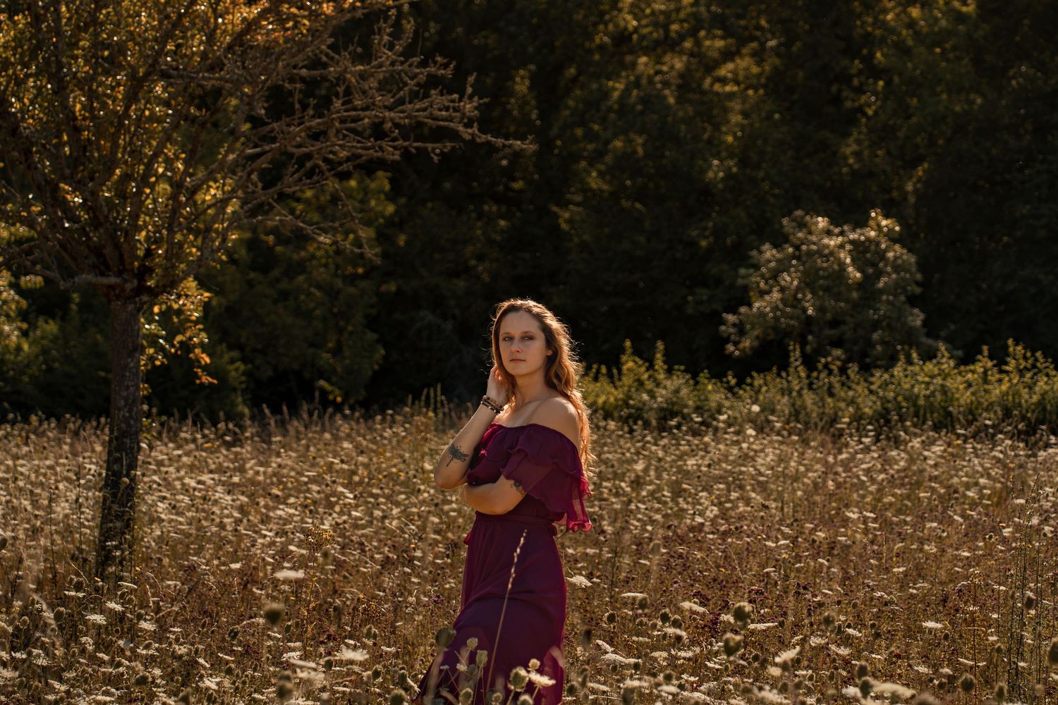 A young woman in a burgundy off-shoulder dress stands in a golden wildflower field with autumn trees behind her.