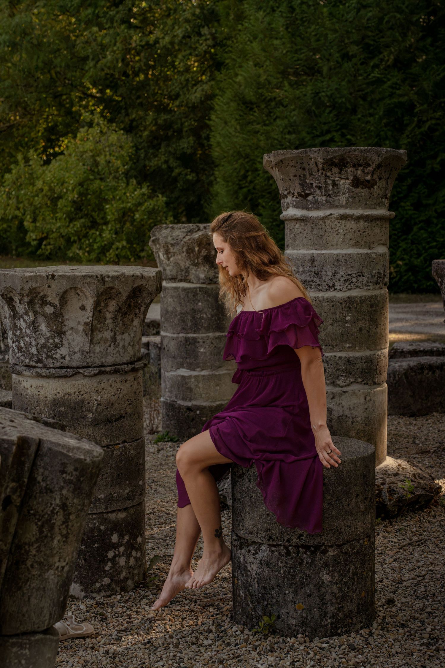 Woman in purple off-shoulder dress sitting on ancient stone column ruins surrounded by lush green trees.