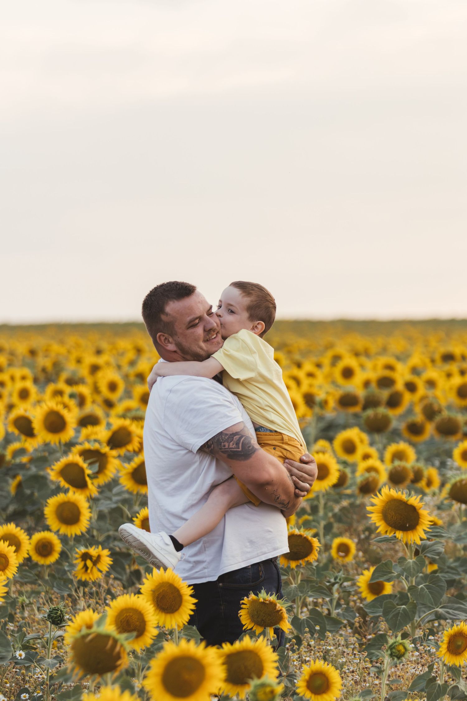 Father holding young son in a vibrant sunflower field, sharing a tender moment with a kiss on the cheek.