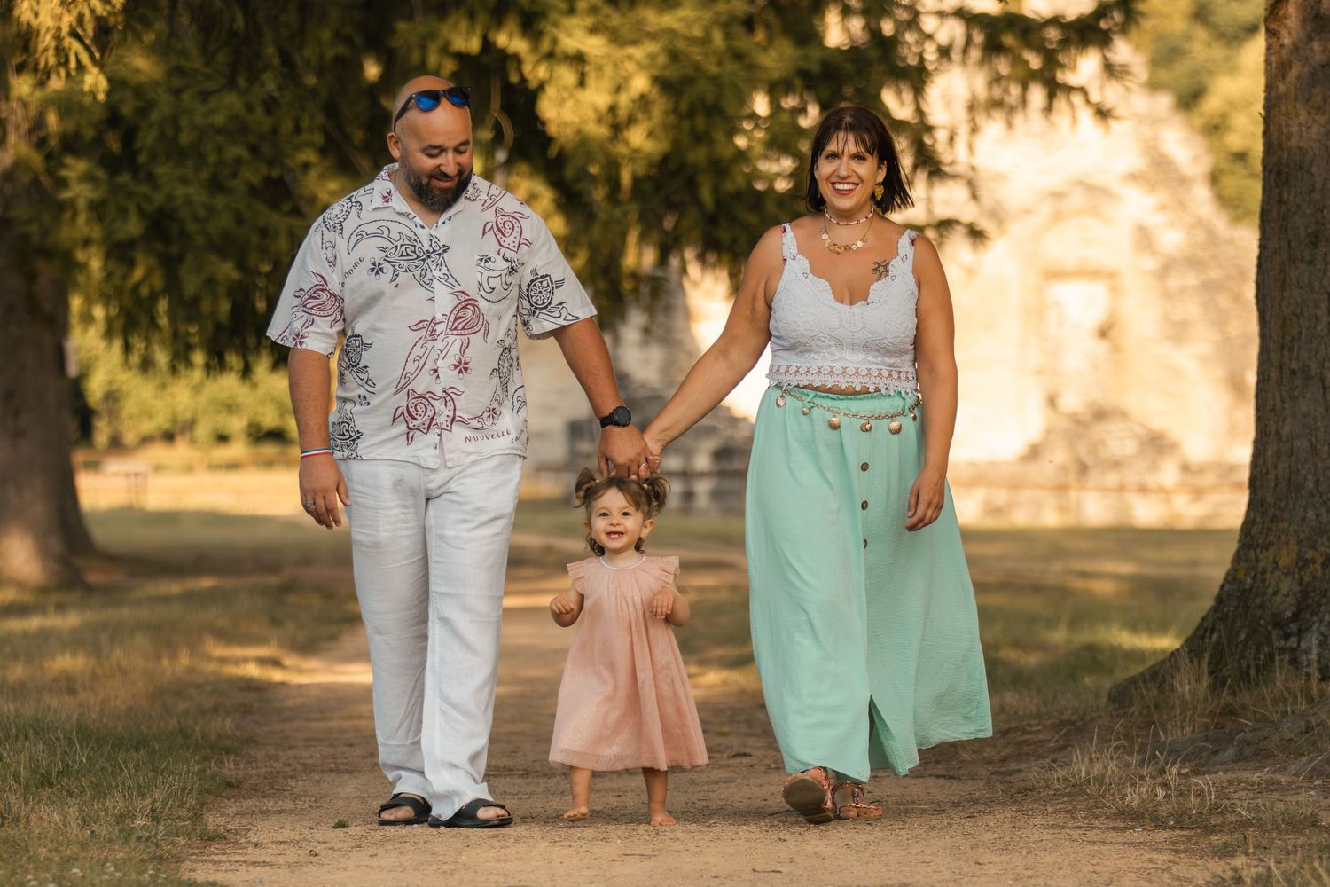 A happy family of three walks hand-in-hand along a sunny tree-lined path, parents guiding their toddler girl in a pink dress.