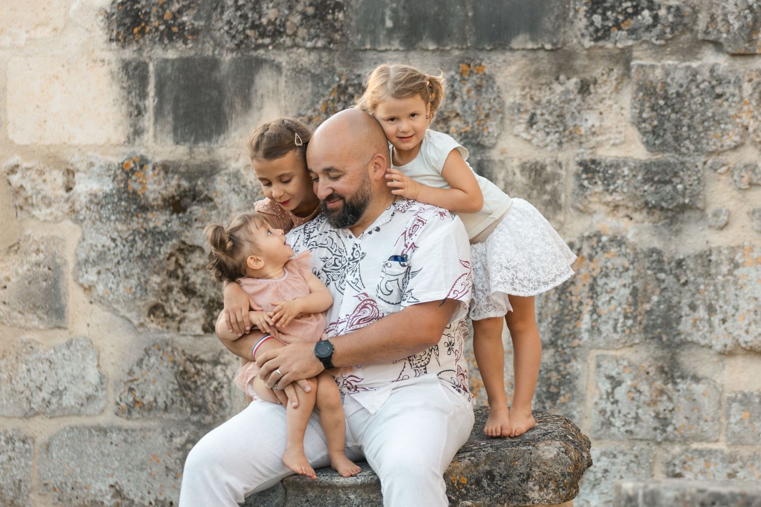 Bald man sitting against stone wall laughing with three young girls climbing on him outdoors.