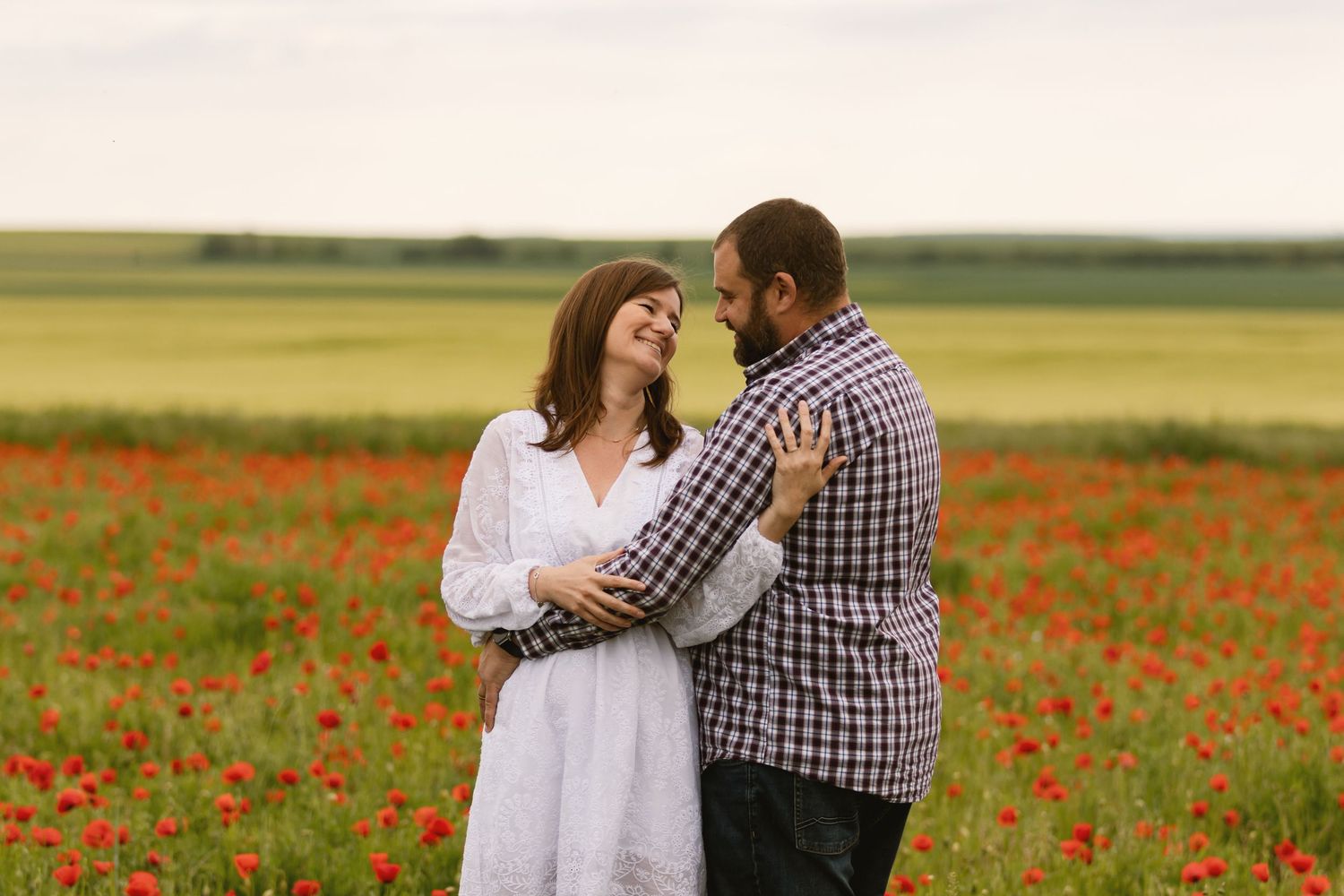 Couple embracing romantically in a vibrant red poppy field, woman in white dress, man in plaid shirt.