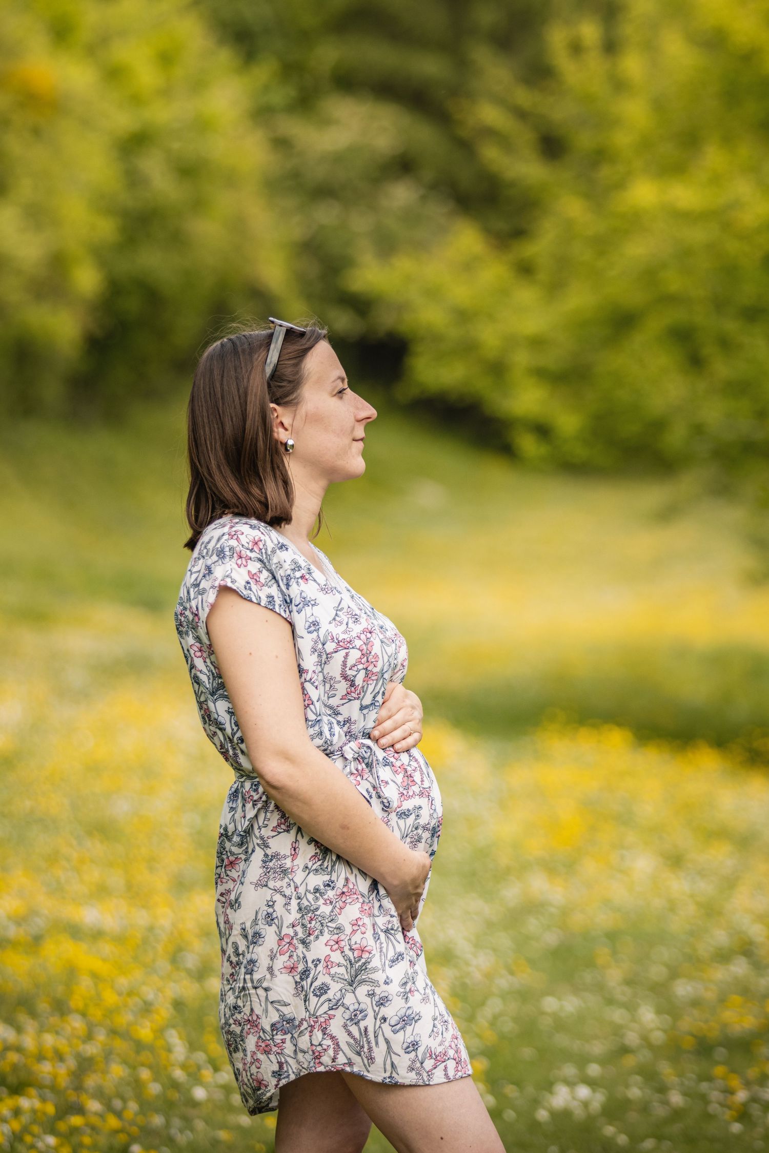 Pregnant woman in floral dress cradling her baby bump in a sunny meadow with wildflowers and green trees.