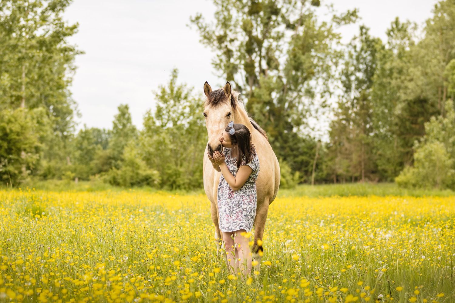 Girl in floral dress hugging a palomino horse in a sunny meadow filled with yellow wildflowers and green trees.