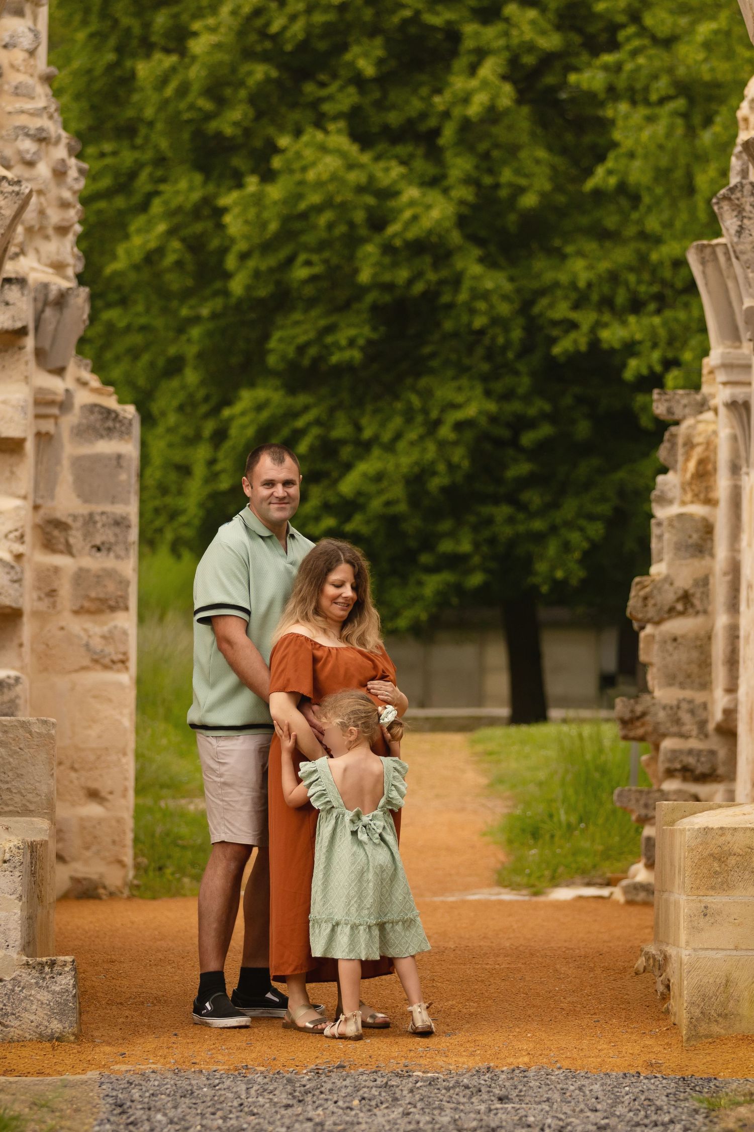 Family of three poses between stone columns in a lush green park, pregnant mother in orange dress with husband and young daughter.
