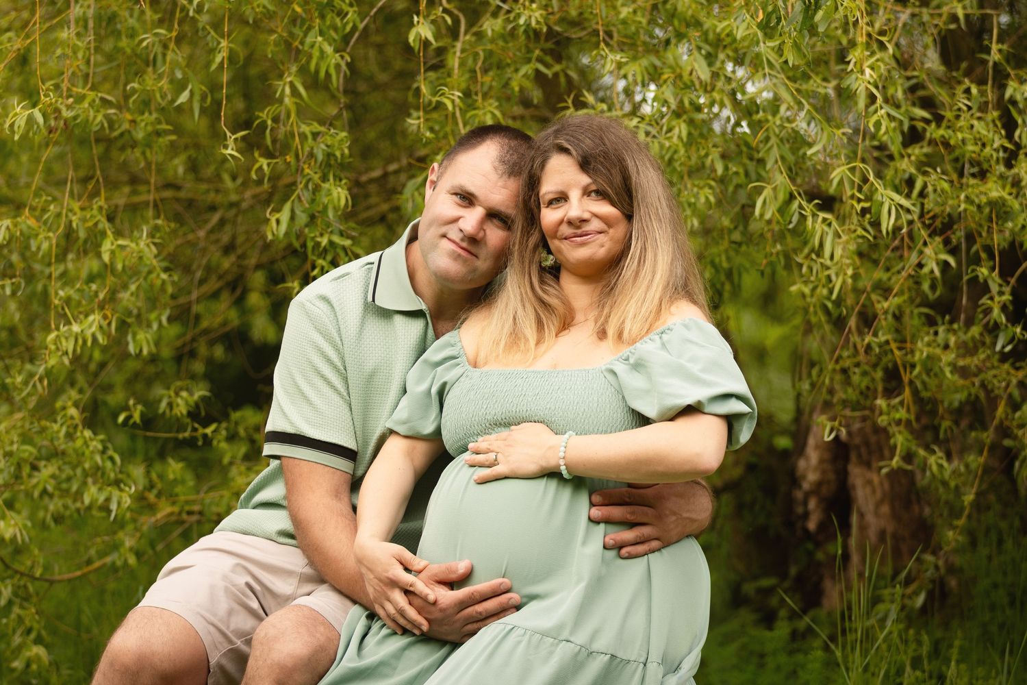 Happy couple in matching green outfits posing outdoors, pregnant woman cradling baby bump near lush green trees.