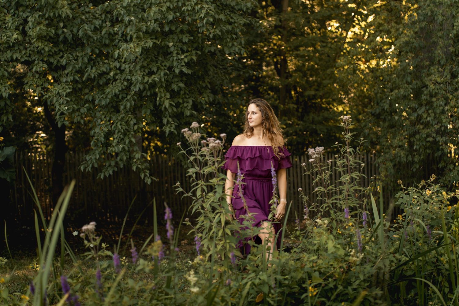 Woman in purple off-shoulder dress standing among lush green garden plants and trees in golden hour light.