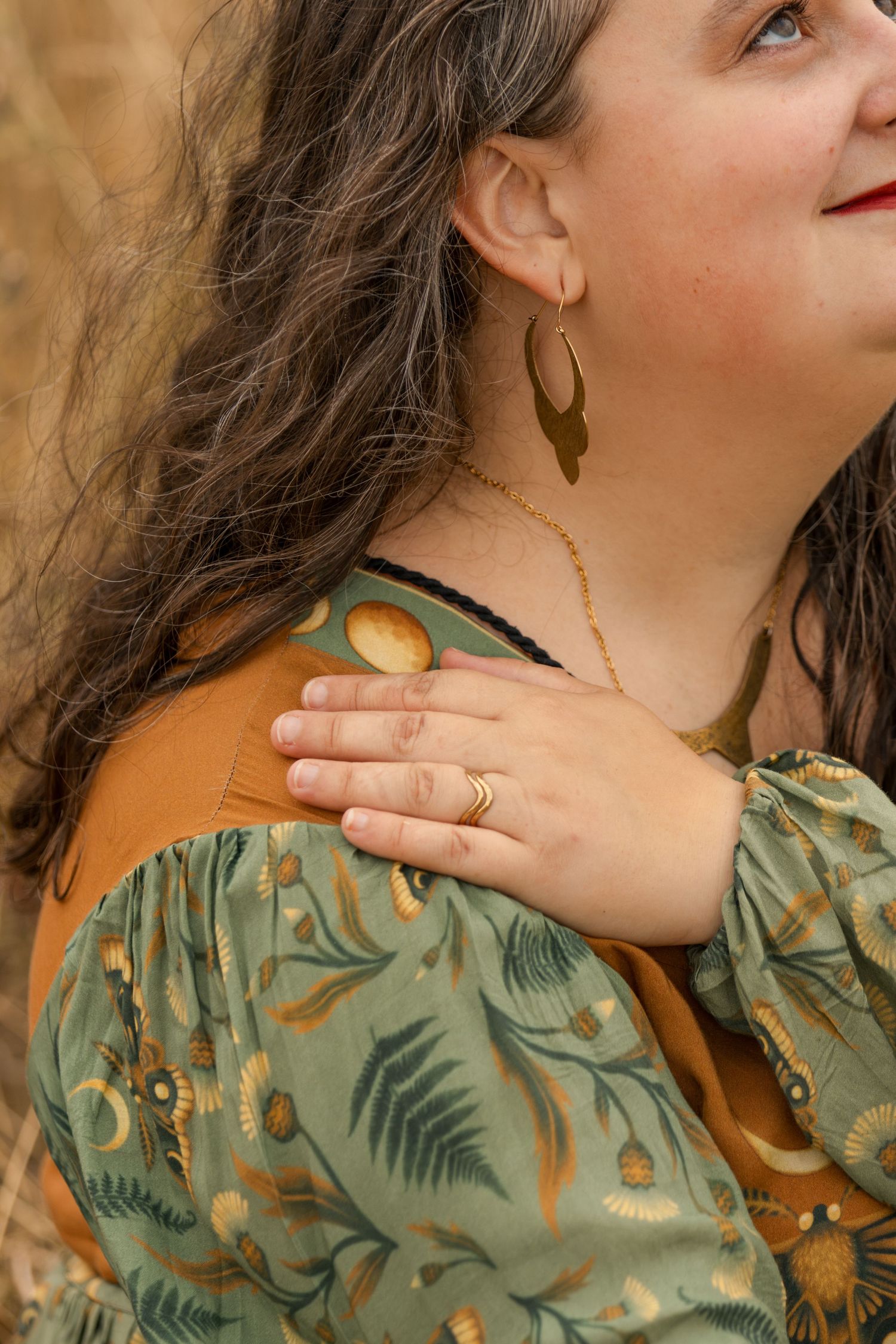 Close-up of woman wearing gold leaf earrings and layered necklaces in a floral dress outdoors.