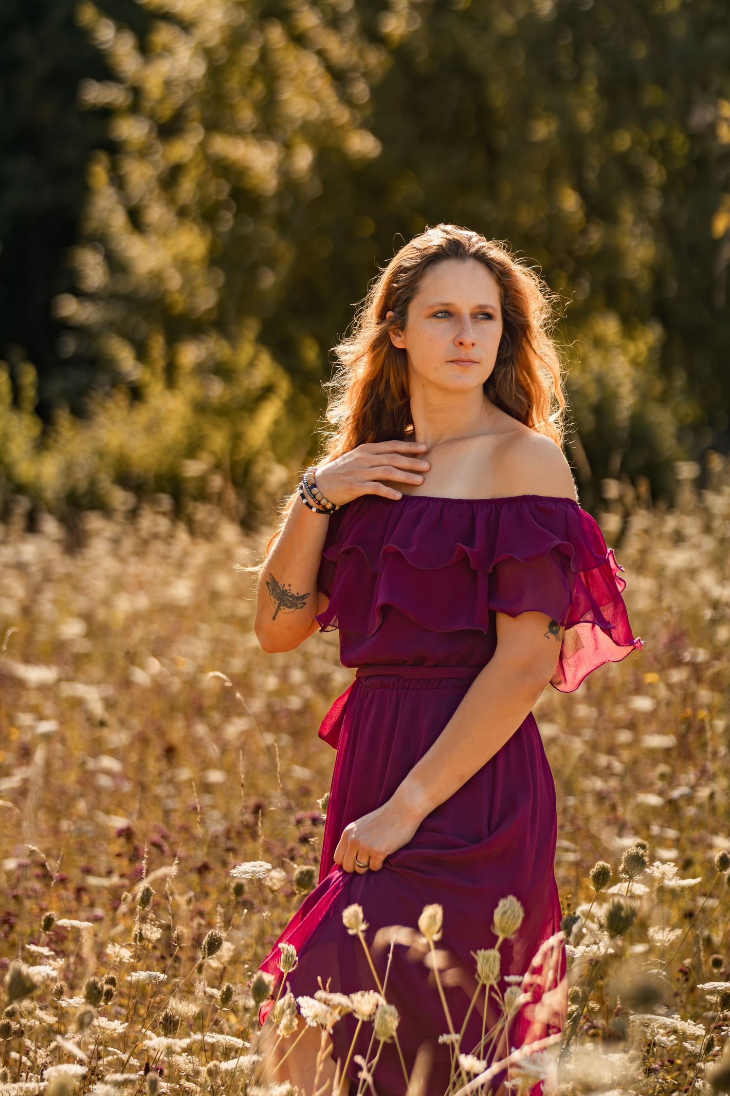 Young woman in burgundy off-shoulder dress standing in golden field with wildflowers, bathed in warm sunlight.