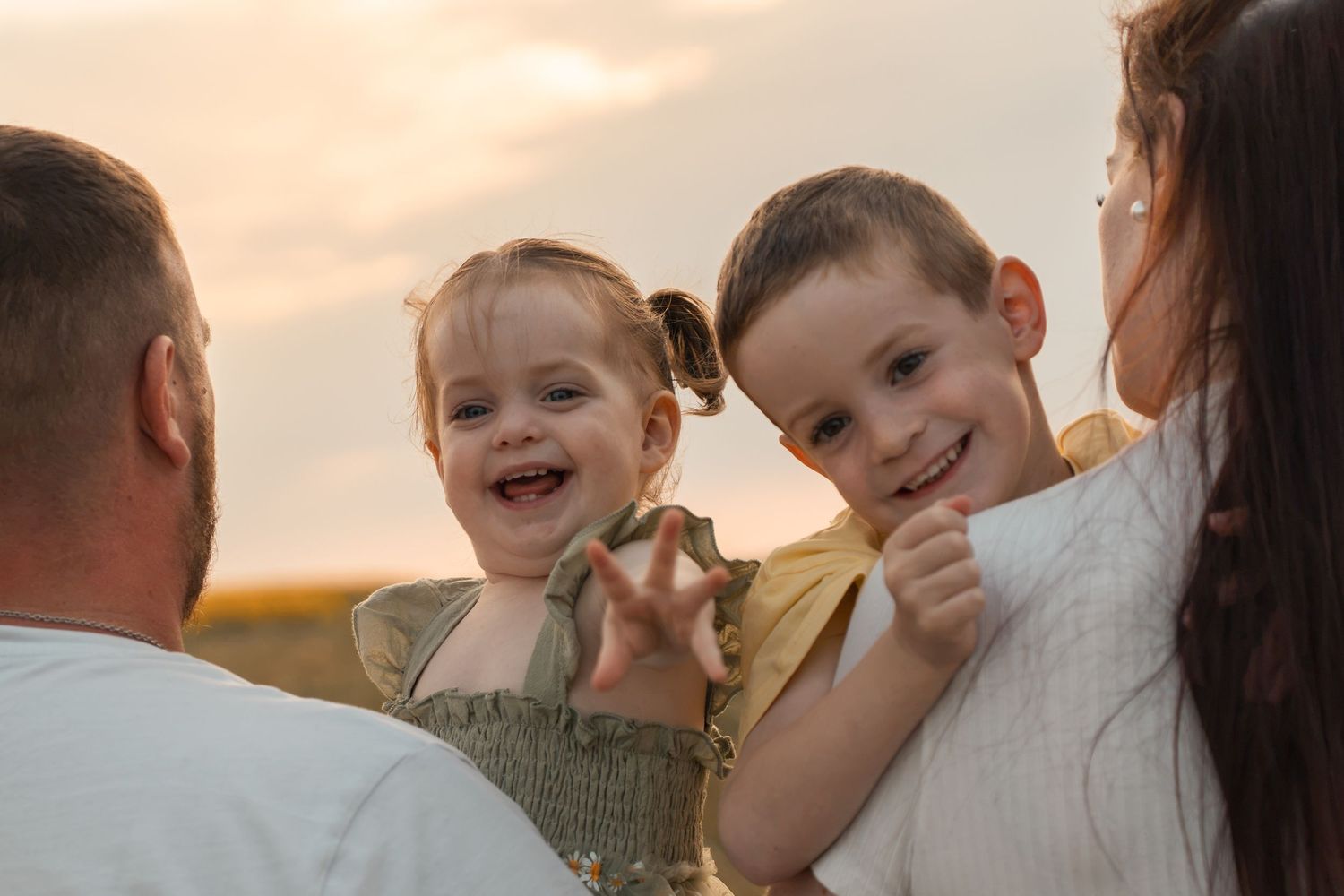 Two happy children smile while being held by their parents outdoors at sunset in a golden field.