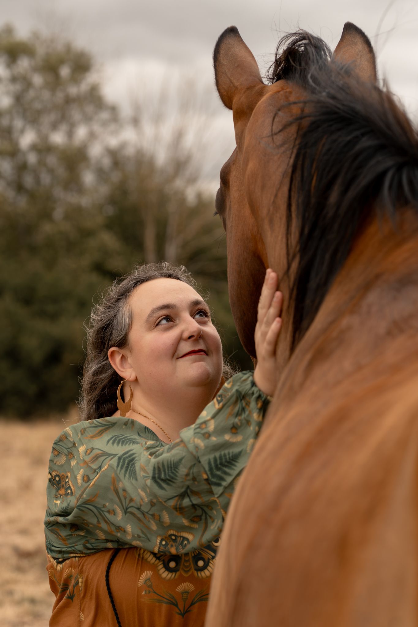 A woman gently touches a brown horse's face outdoors, sharing a tender moment with the animal against a cloudy sky.