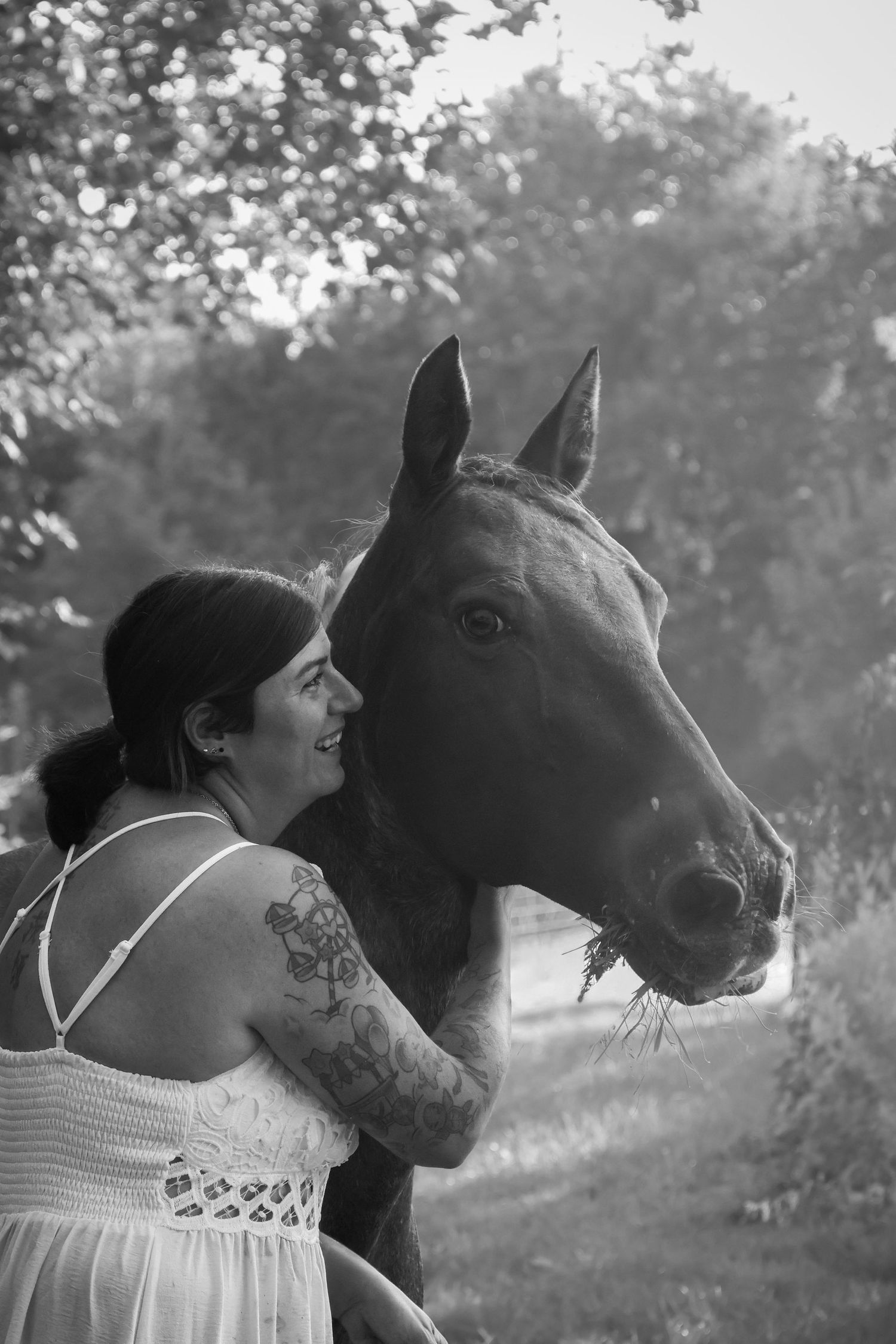 Black and white image of a laughing tattooed woman standing next to a dark horse.