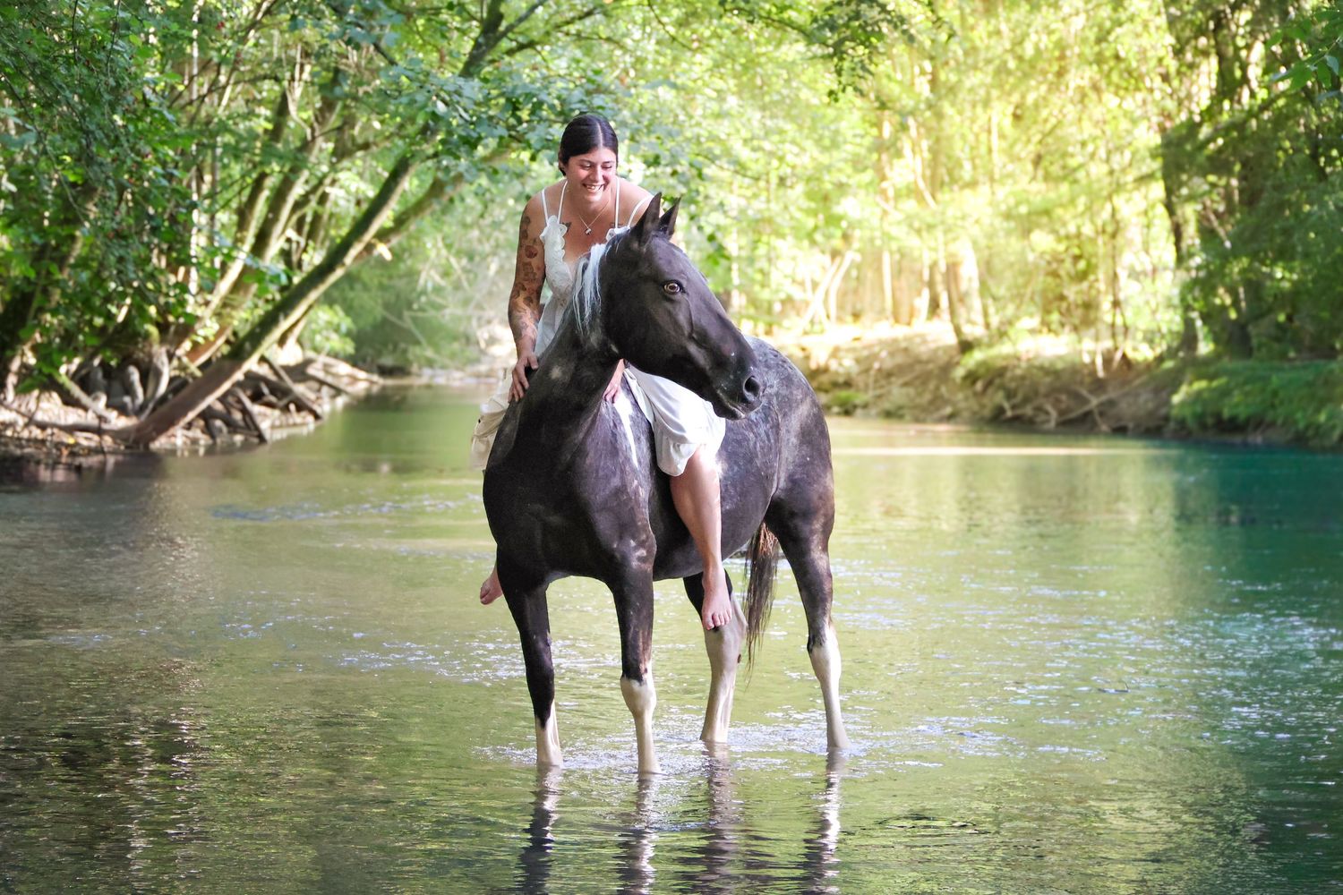 Woman leaning forward on a dark horse wading through a peaceful forest creek.