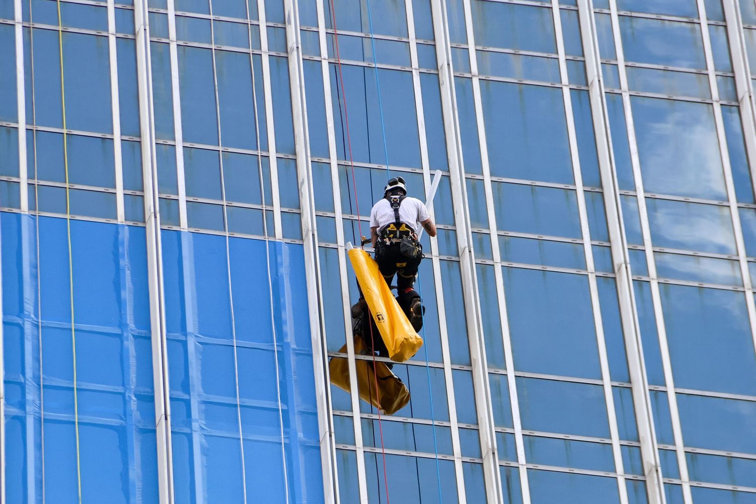 Window washer on yellow scaffold cleaning blue glass skyscraper facade on a sunny day.