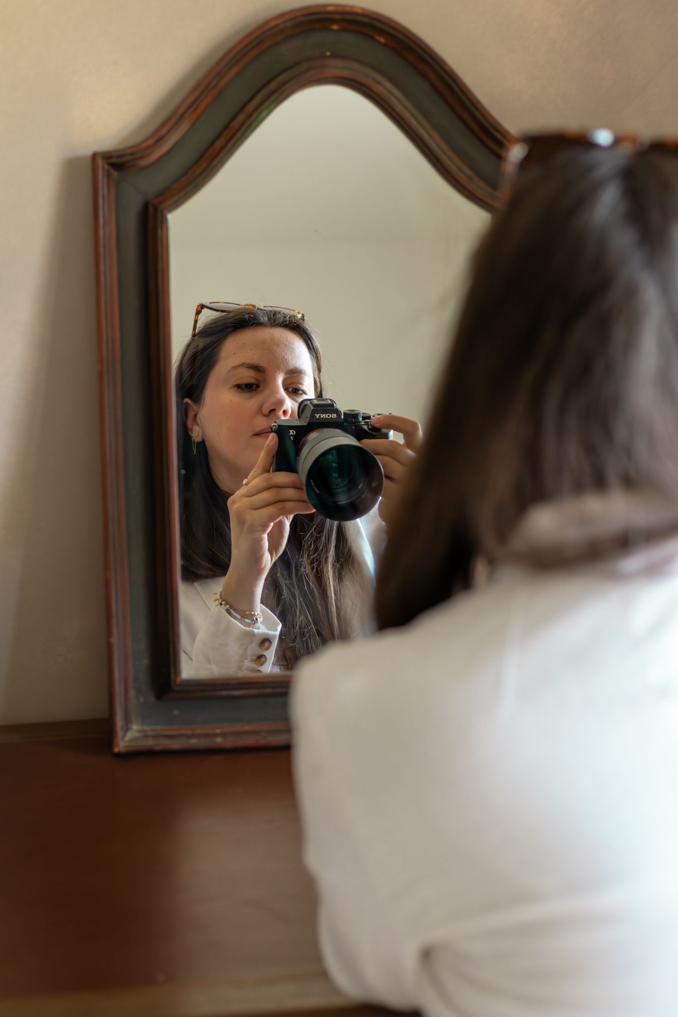 Young woman photographing herself in an ornate wooden mirror using a Nikon DSLR camera, wearing a white blazer.