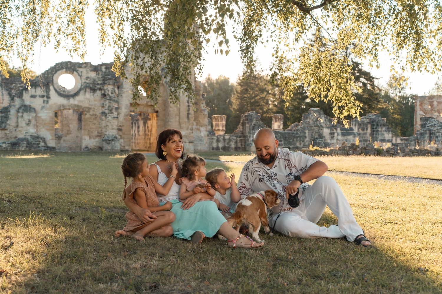 A happy family with children and a dog relaxes on grass near historic ruins on a sunny day.