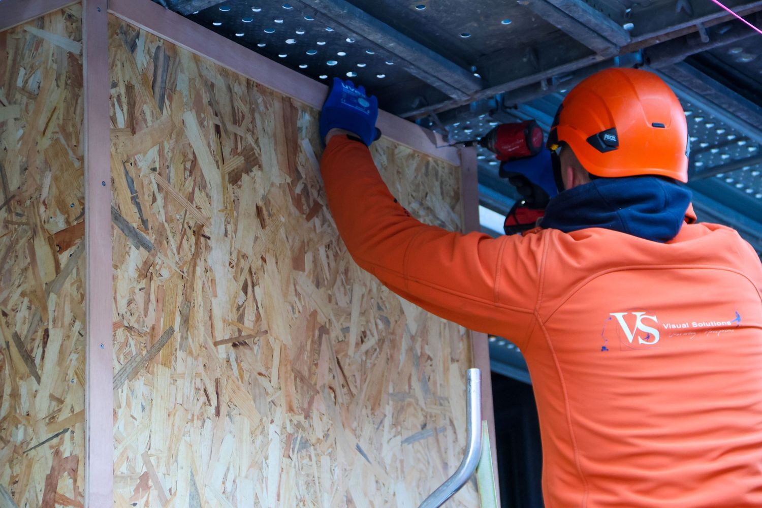Worker in orange uniform and red helmet installing OSB board panel on a wall frame at a construction site.