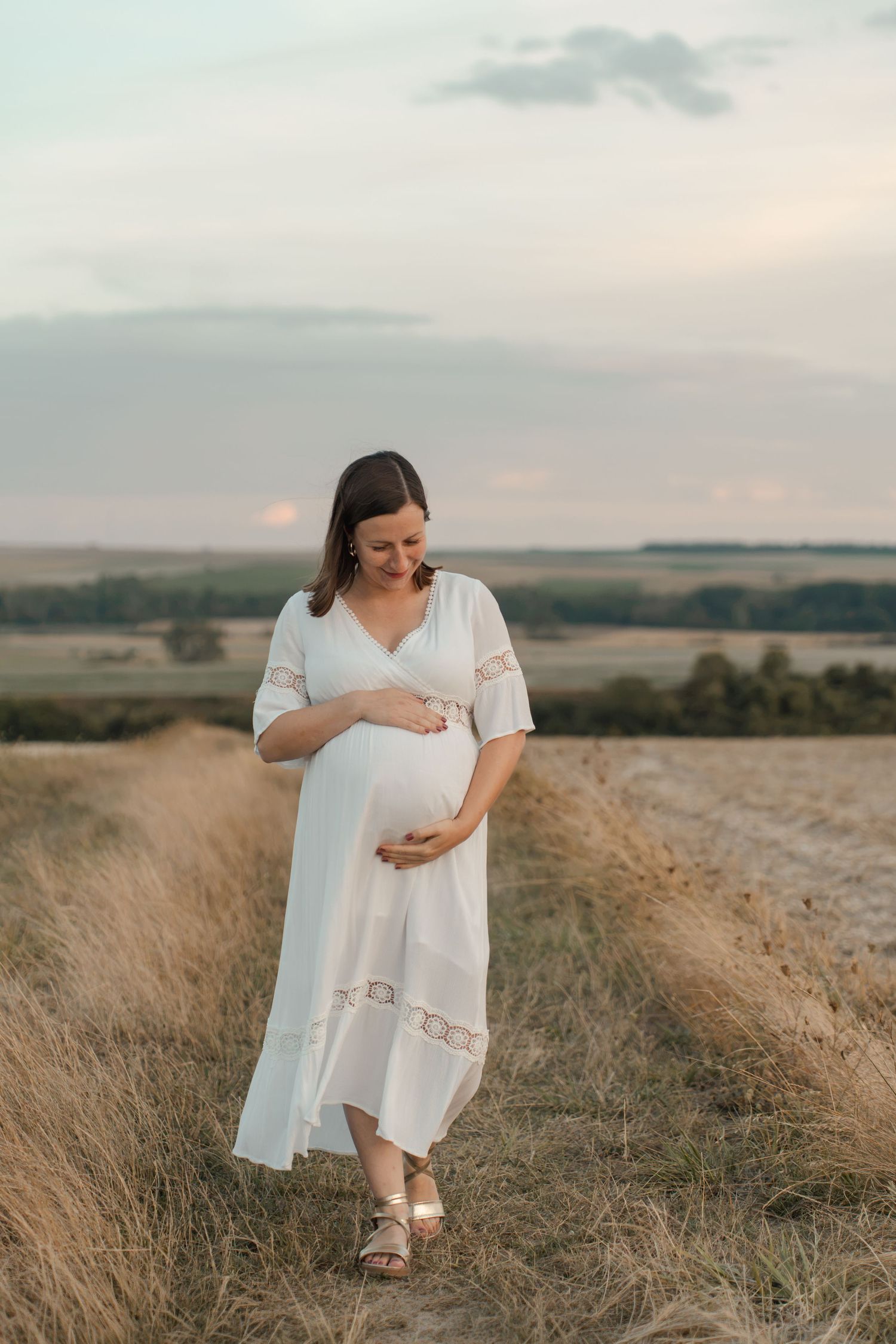 Pregnant woman in white lace dress standing in golden field at sunset, cradling her baby bump.