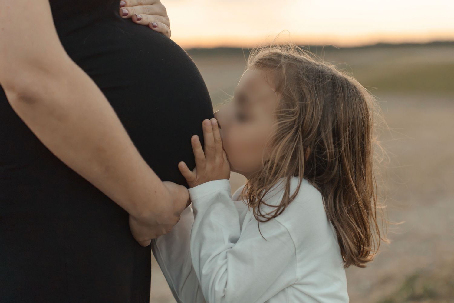 Young girl kissing pregnant mother's belly outdoors at sunset in a tender maternity photography moment.