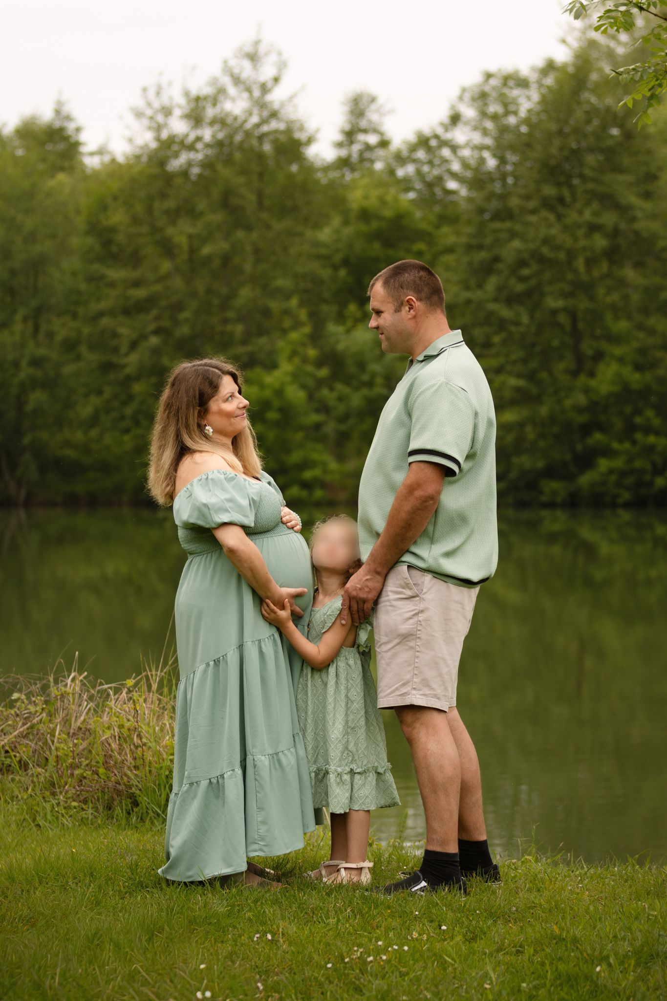 Pregnant woman in mint dress with husband and young daughter standing by a serene pond surrounded by lush green trees.