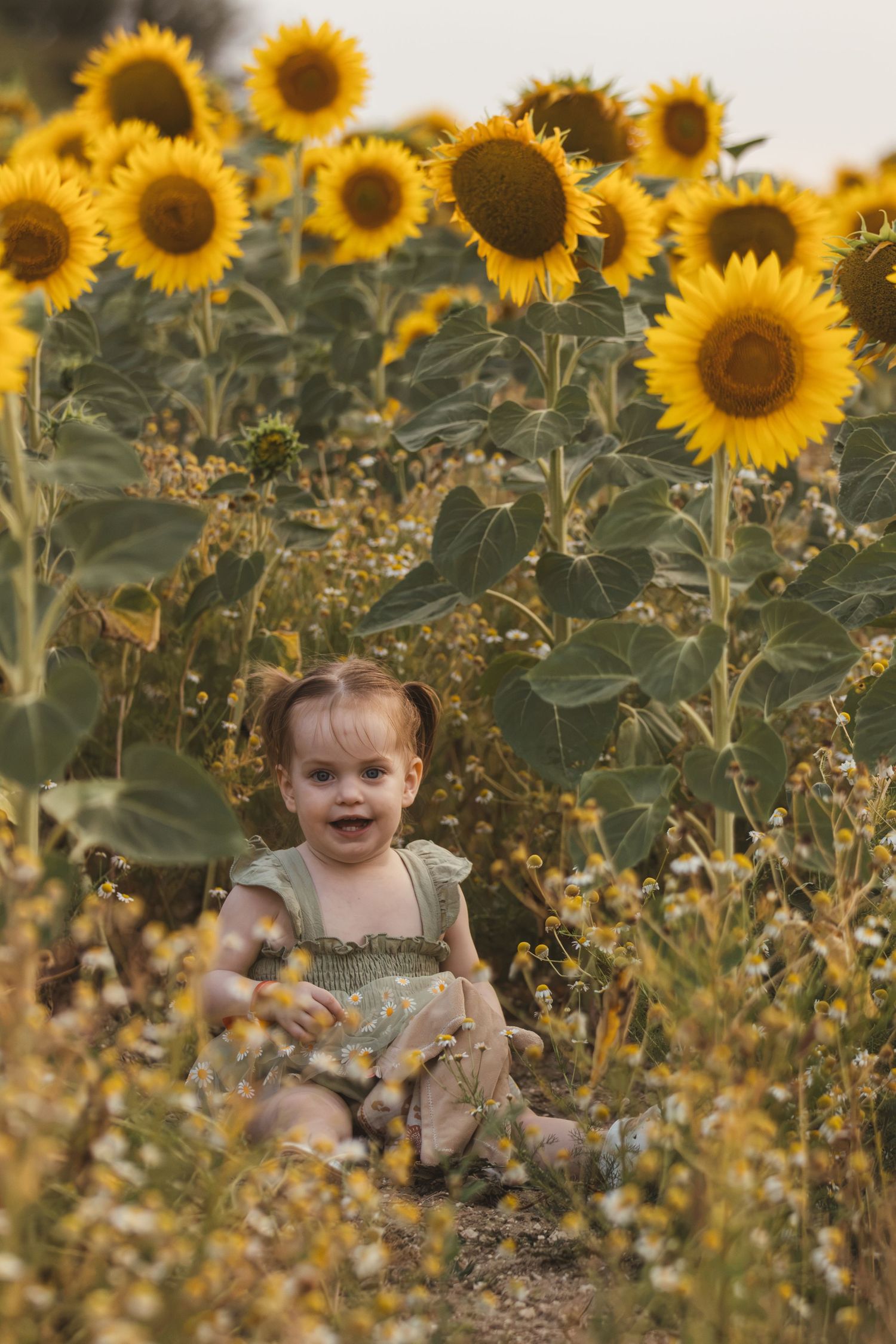 Smiling toddler girl in green dress sitting in a vibrant sunflower field on a sunny day.