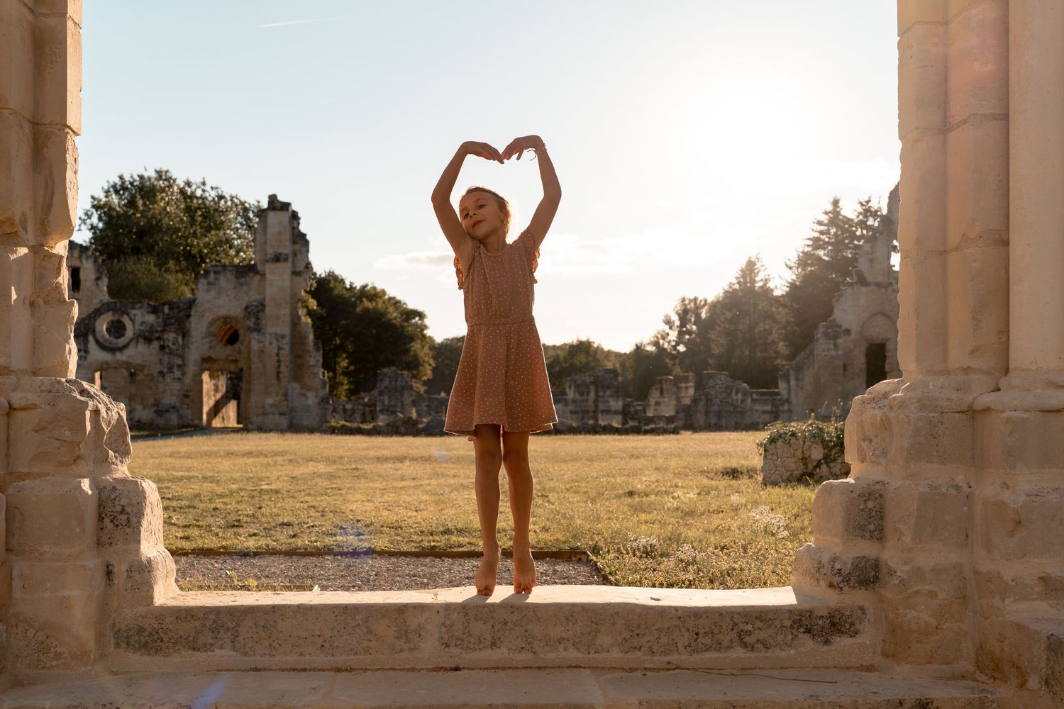 Woman in beige dress making heart shape with arms at ancient ruins in sunlight.