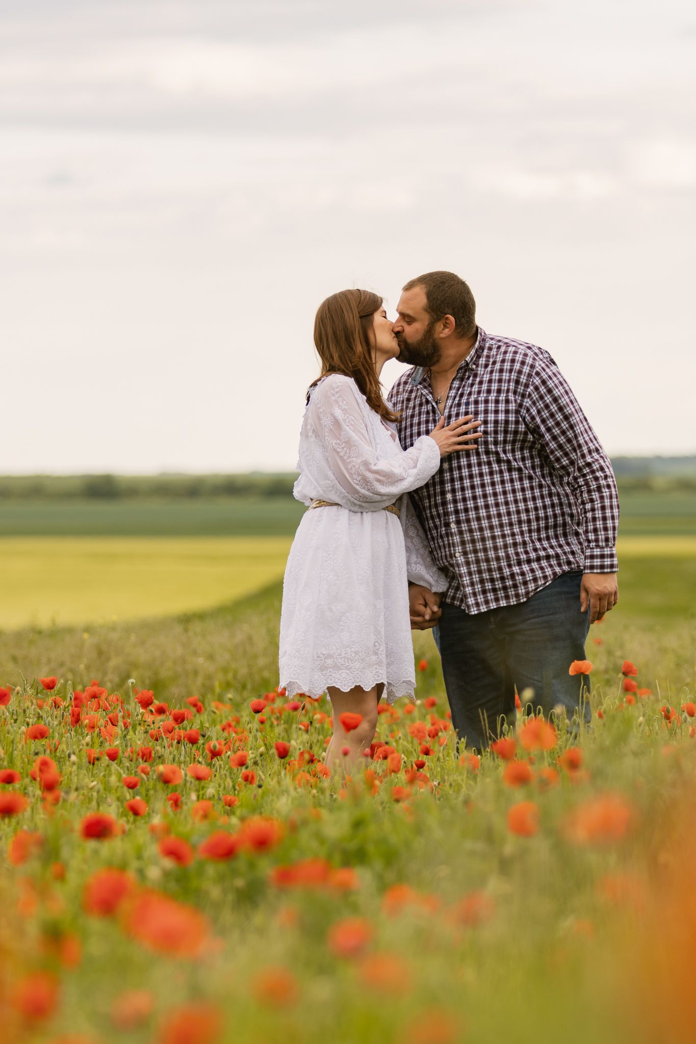 Couple sharing a romantic kiss in a vibrant red poppy field, woman in white dress, man in plaid shirt.