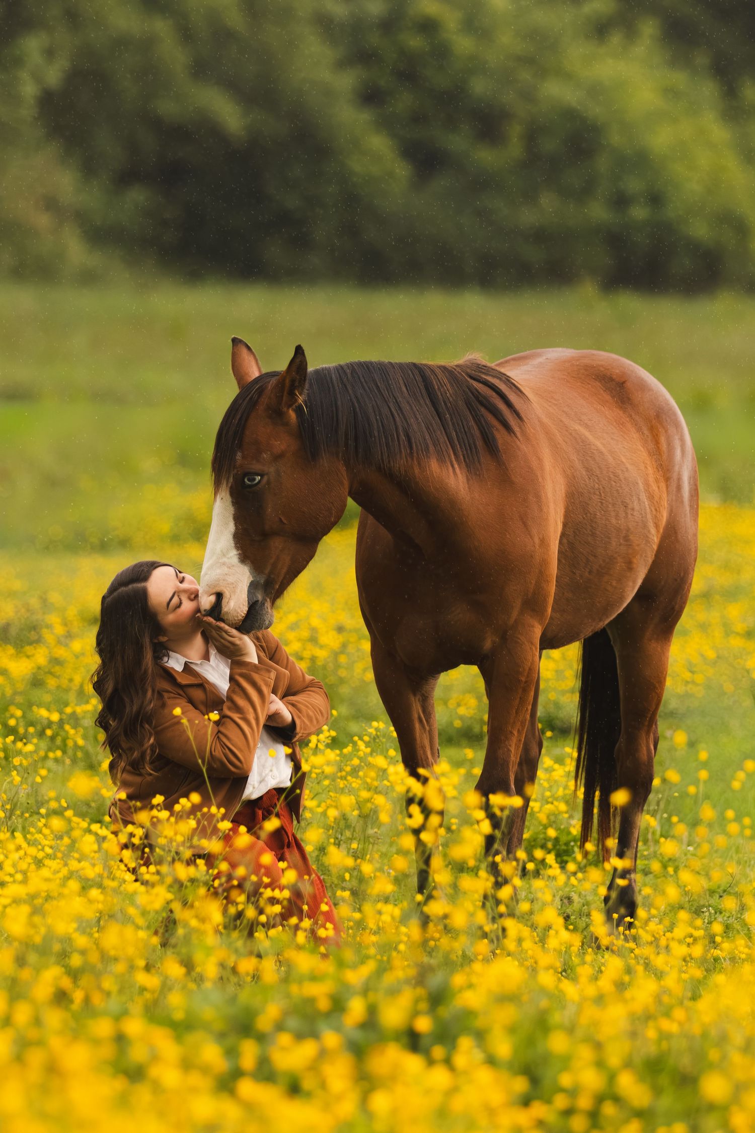 Woman in brown jacket sits among yellow wildflowers with a chestnut horse nuzzling her in a green field.