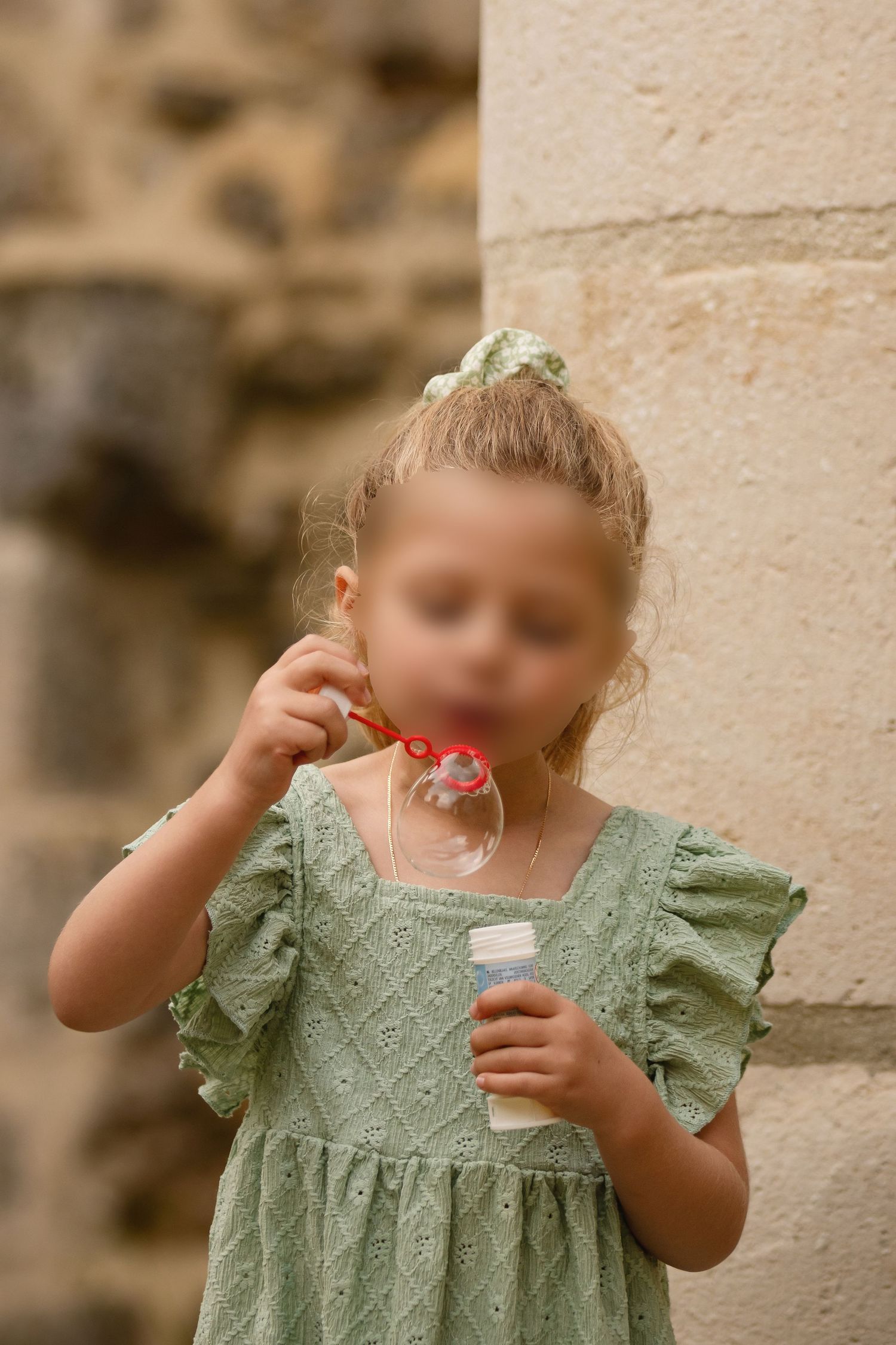 Young girl in green dress blowing soap bubbles outdoors near stone wall.