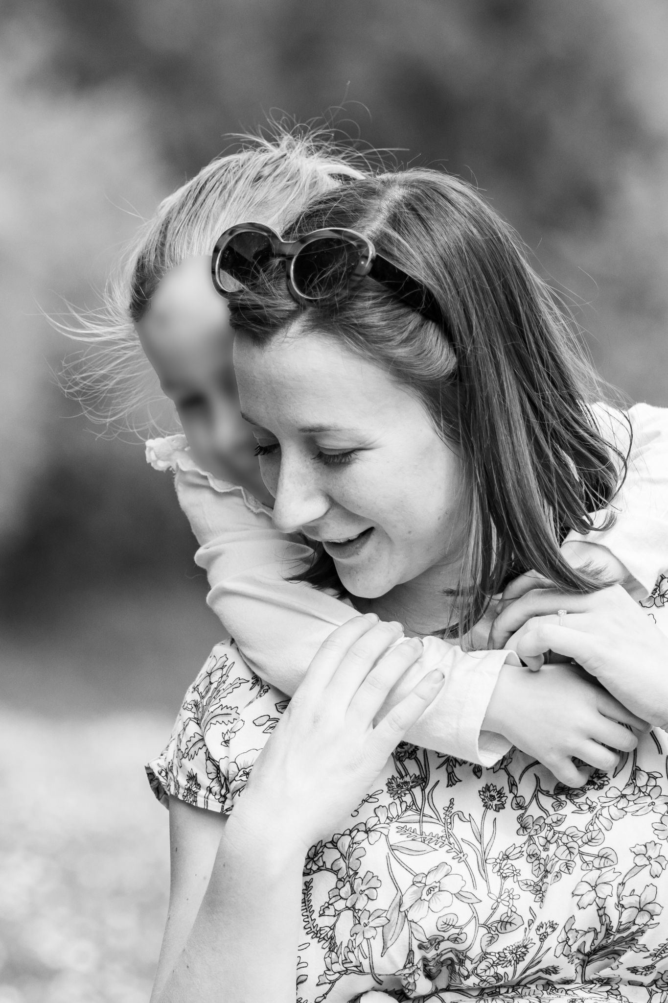 Black and white photo of a laughing woman being hugged from behind by a child outdoors.