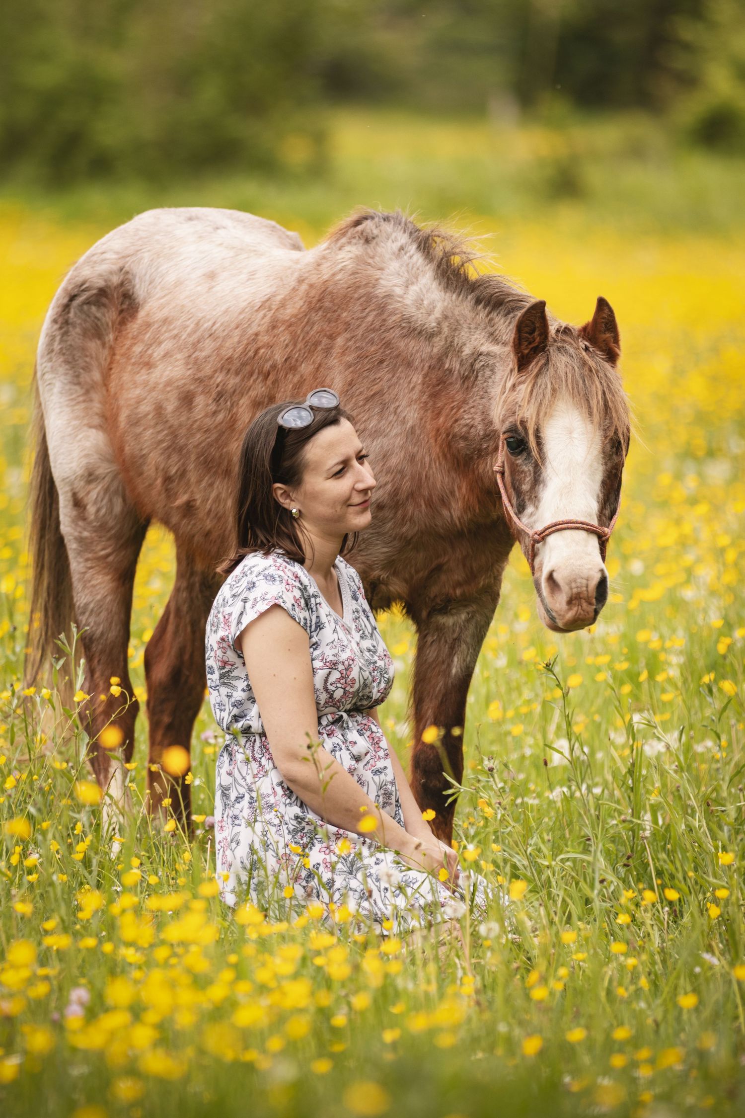 Woman in floral dress sitting in yellow wildflower field beside a brown roan horse on a sunny day.