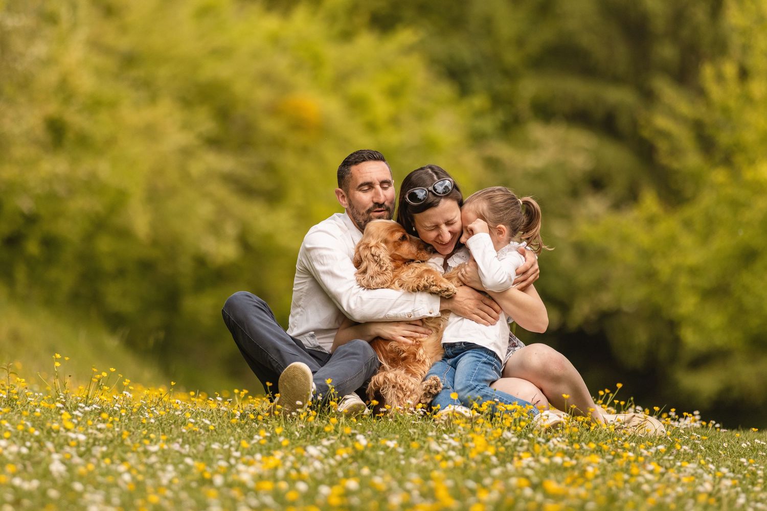 Family with a golden retriever dog sitting together in a sunny meadow with yellow wildflowers and green trees.