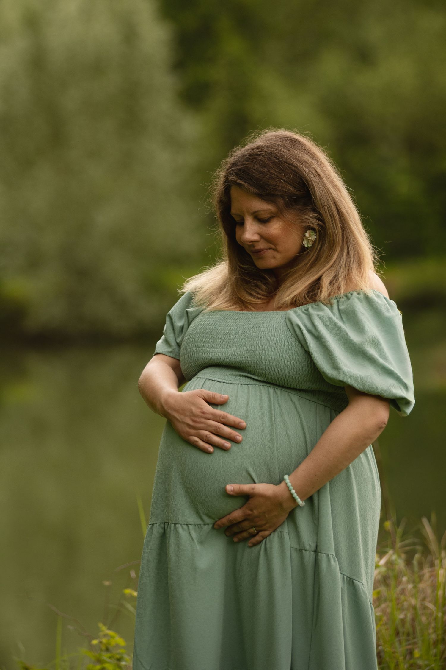 Pregnant woman in green dress cradling her baby bump outdoors near a serene lake surrounded by lush greenery.