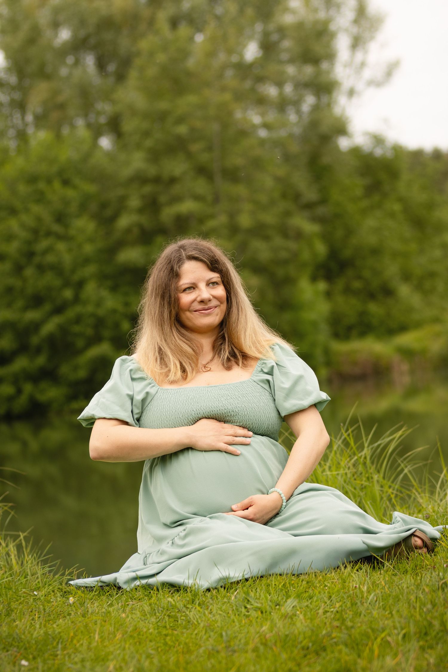 Pregnant woman in sage green dress sitting on grass by a pond, smiling and cradling her baby bump outdoors.