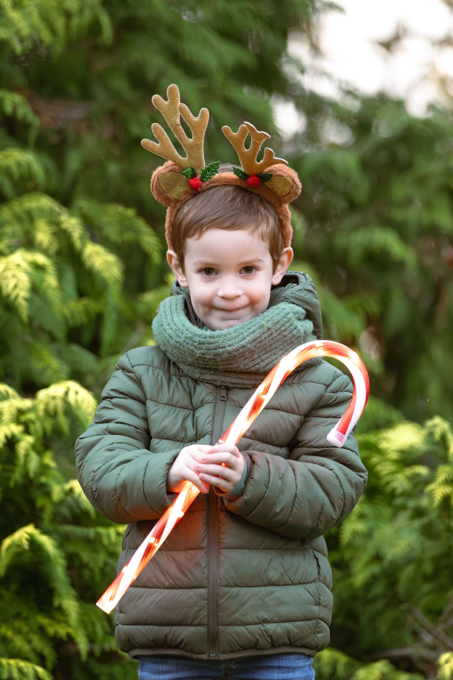 A smiling boy in a green jacket wears reindeer antlers and holds a large candy cane.